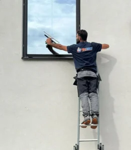 En man klädd i mörka arbetskläder, med en logotyp på ryggen som delvis döljs av armen, står på en metallstege lutad mot en vit husvägg. Han sträcker sig för att putsa ett fönster med en fönsterskrapa. Huset är modernt och fönstret är mörkt inramat